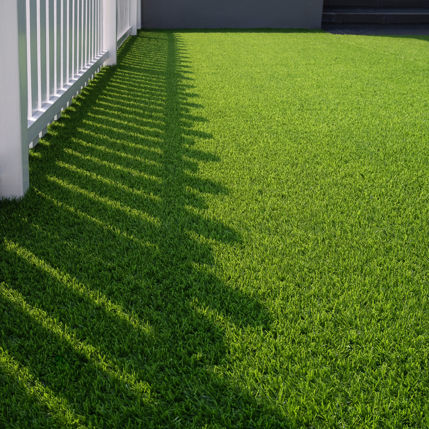 Sunlight and shadow of white wooden fence on green artificial turf surface in front yard of home, selective focus with copy space